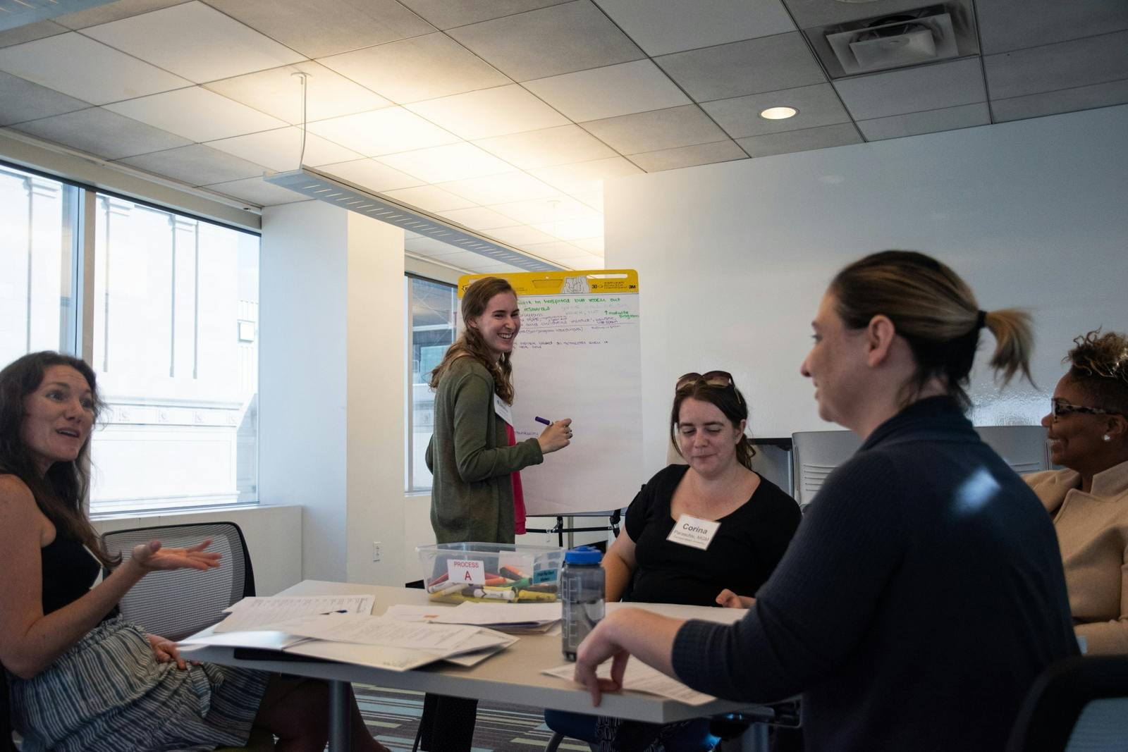 A group of young adult women sit around a table smiling and talking while a woman stands and writes on a white board.