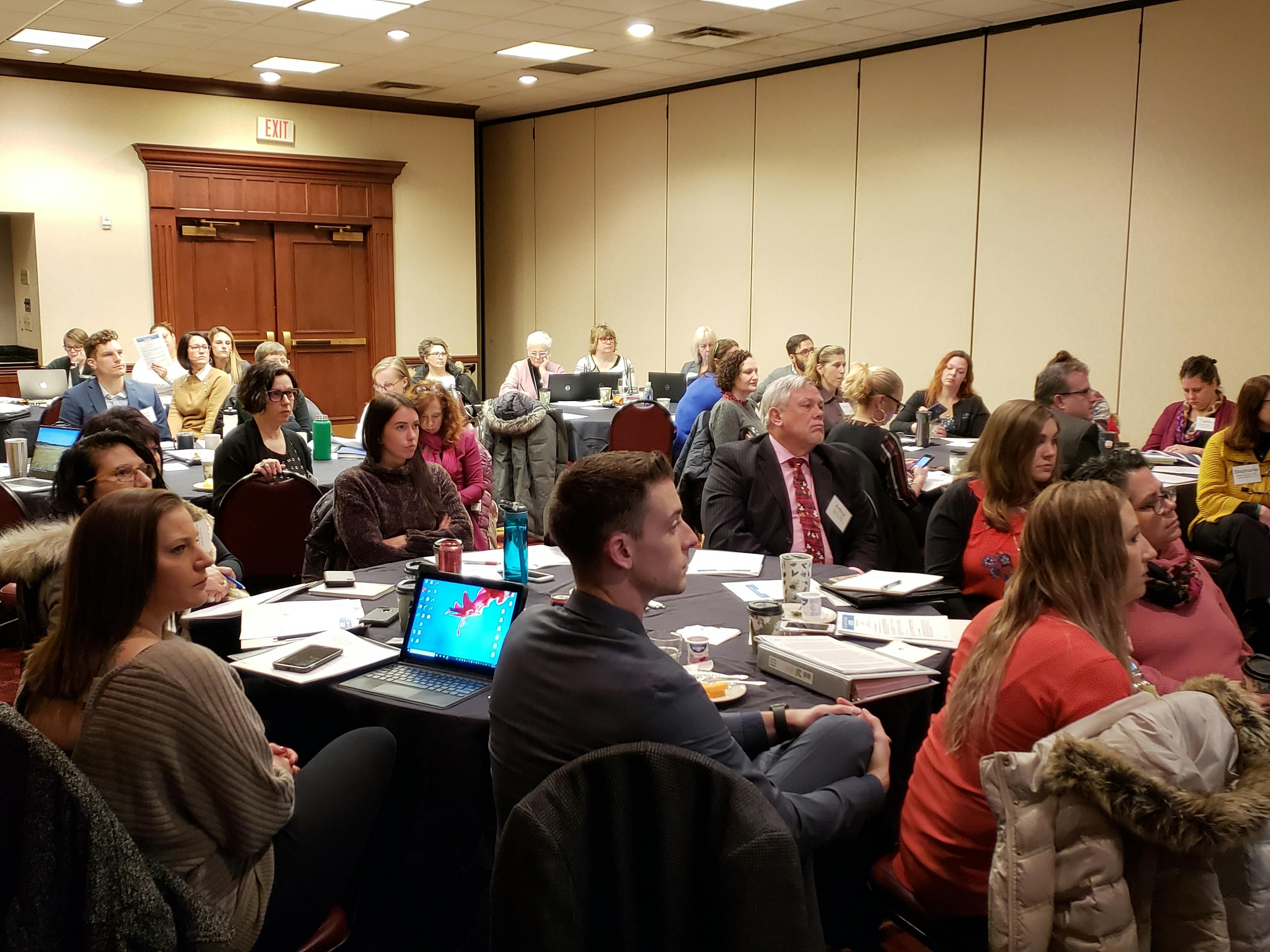 Meeting participants seated around numerous round tables and listening to a speaker