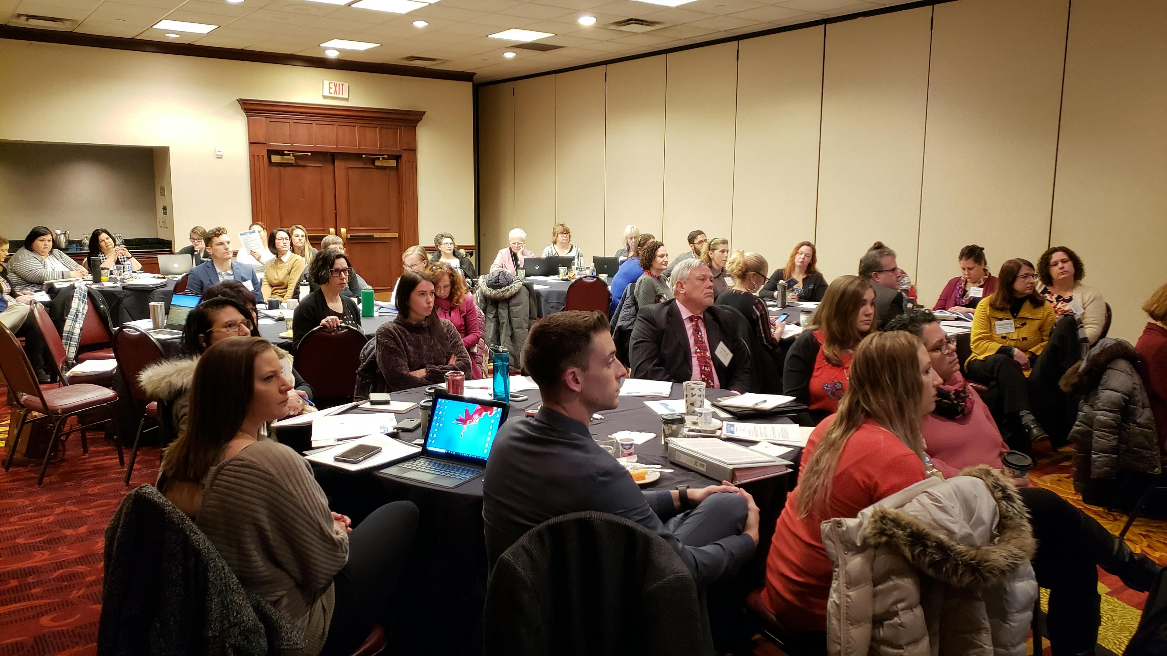 Meeting participants seated around numerous round tables and listening to a speaker