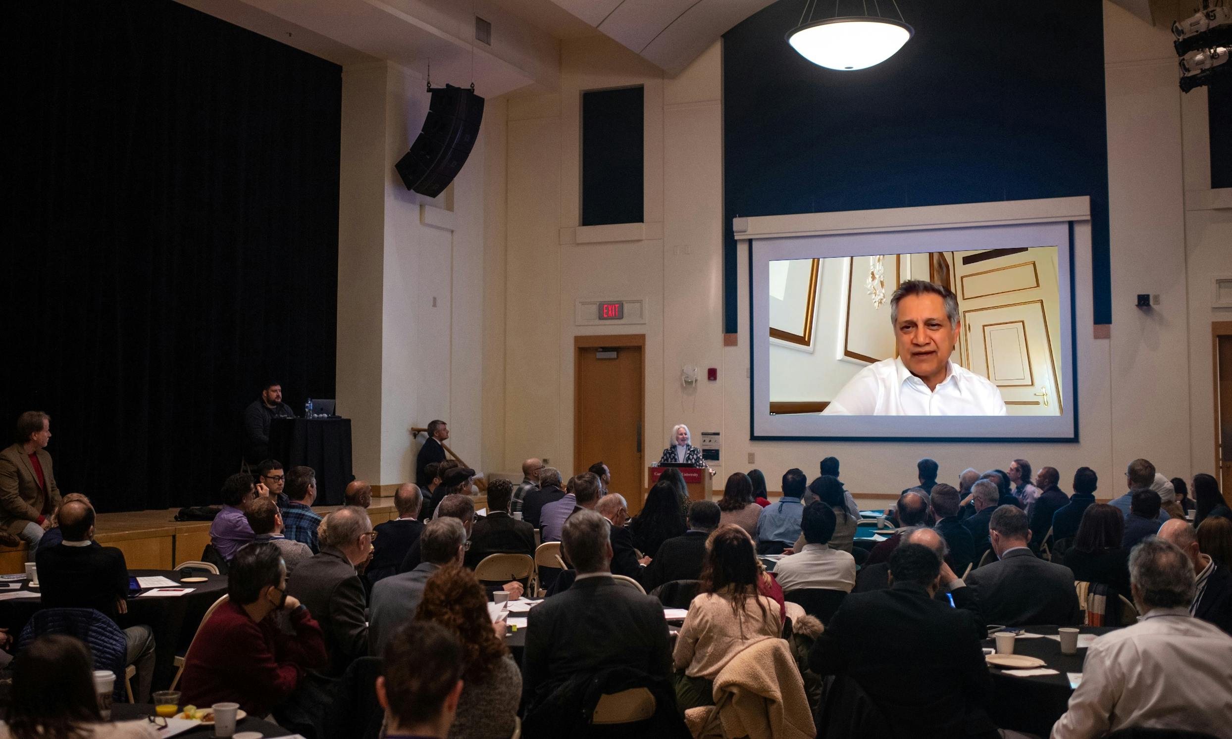 A large room of people sitting at round tables and watching a presentation.